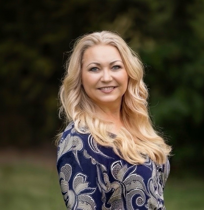 A woman with long, light wavy hair stands in front of a bamboo backdrop, wearing a brown blazer and looking directly at the camera, creating a warm, professional portrait often used for mental‑health therapist profiles and counseling websites